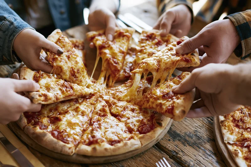 Hands reaching for slices of cheese pizza on a wooden table during a group meal