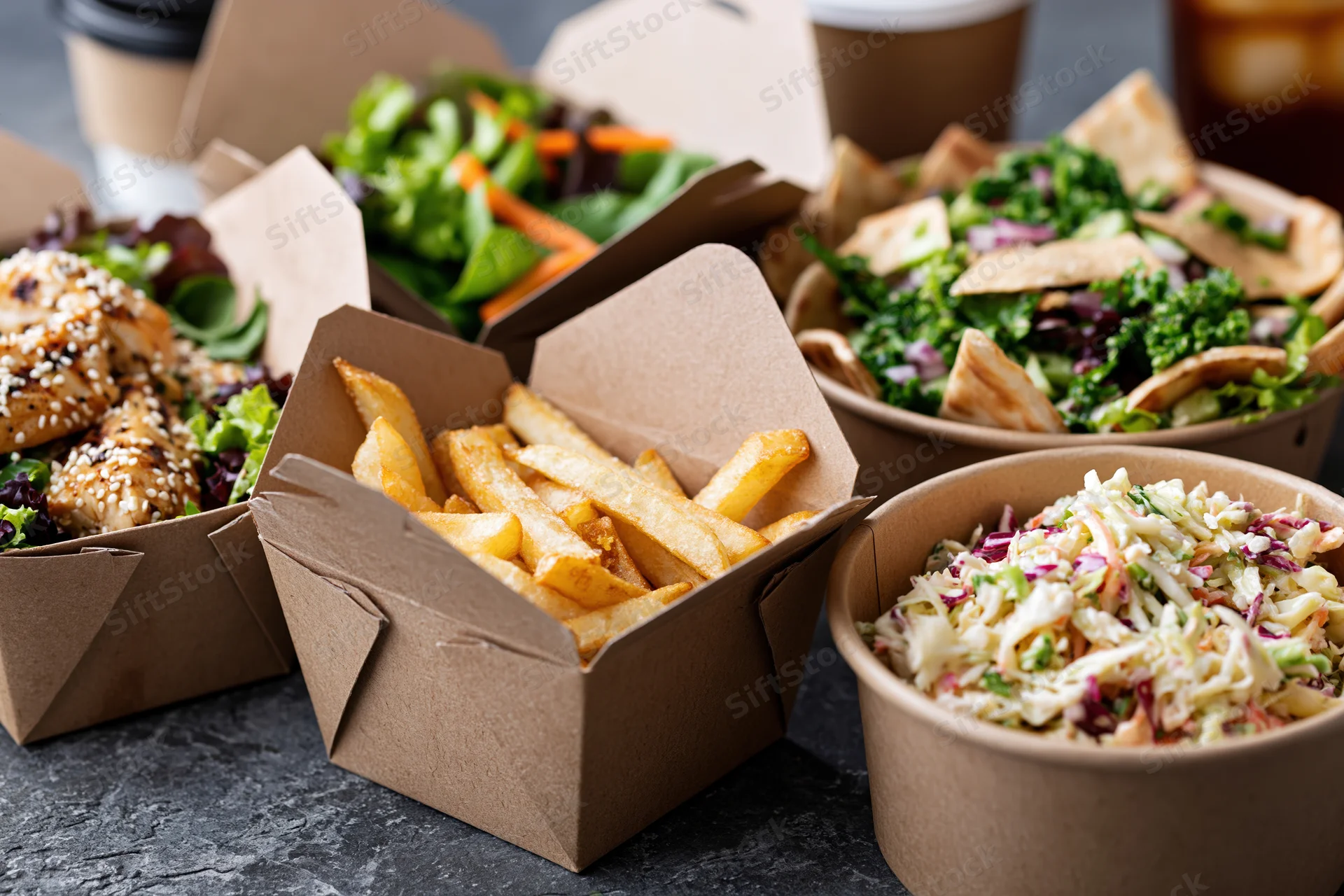 Several kraft paper takeout boxes filled with fries, salads, coleslaw and chicken on a dark surface