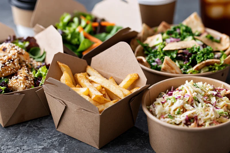 Several kraft paper takeout boxes filled with fries, salads, coleslaw and chicken on a dark surface