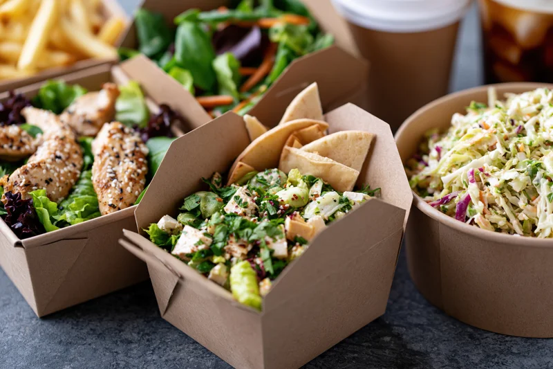 Several kraft paper takeout boxes filled with salads, chicken, fries, and coleslaw arranged on a gray surface