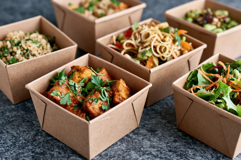 Several kraft paper takeout containers filled with prepared meals arranged on a dark surface