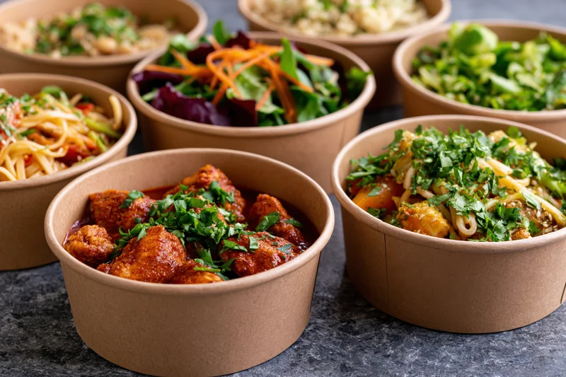 Several kraft paper takeout containers filled with various prepared meals arranged on a dark surface