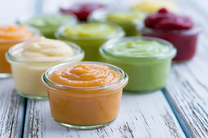 Small glass jars filled with colorful baby food puree arranged on a white wooden table