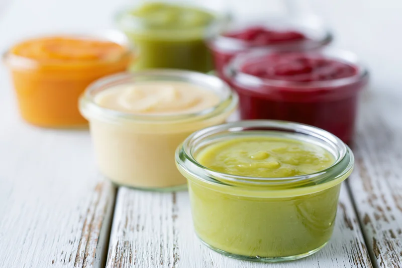 Several small open glass jars filled with colorful baby food puree arranged on a white wooden table