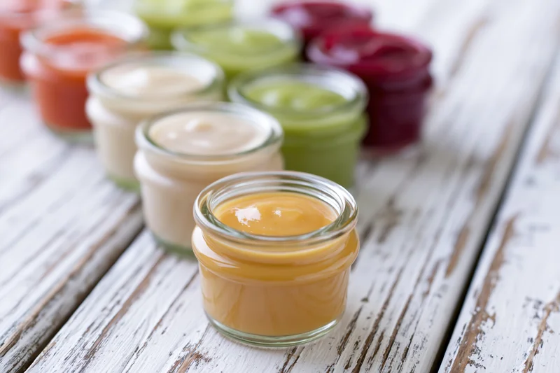 Small glass jars filled with colorful baby food puree arranged in rows on a white distressed wooden table