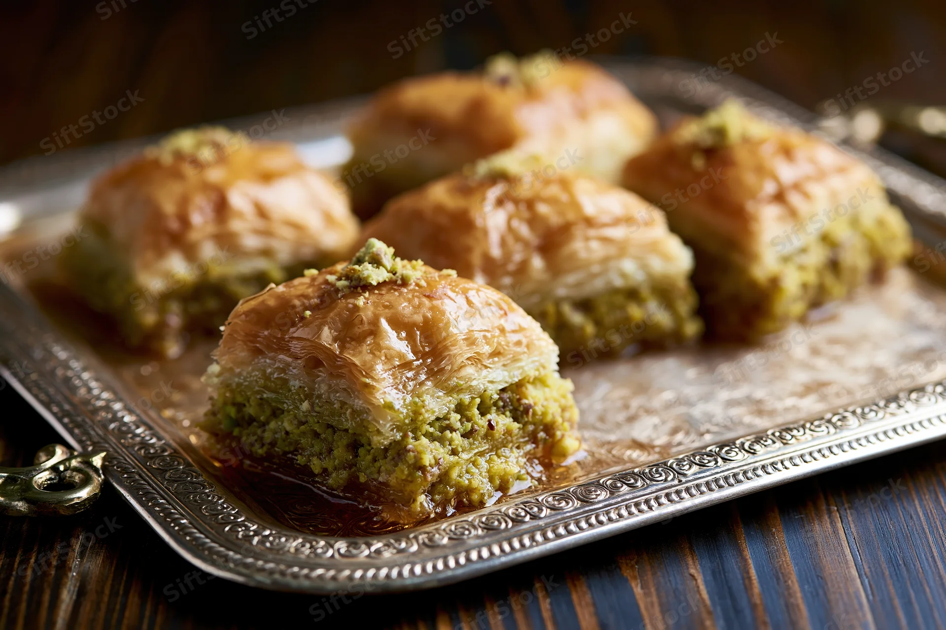 Square pistachio baklava pieces arranged on an ornate silver tray on a dark wooden table