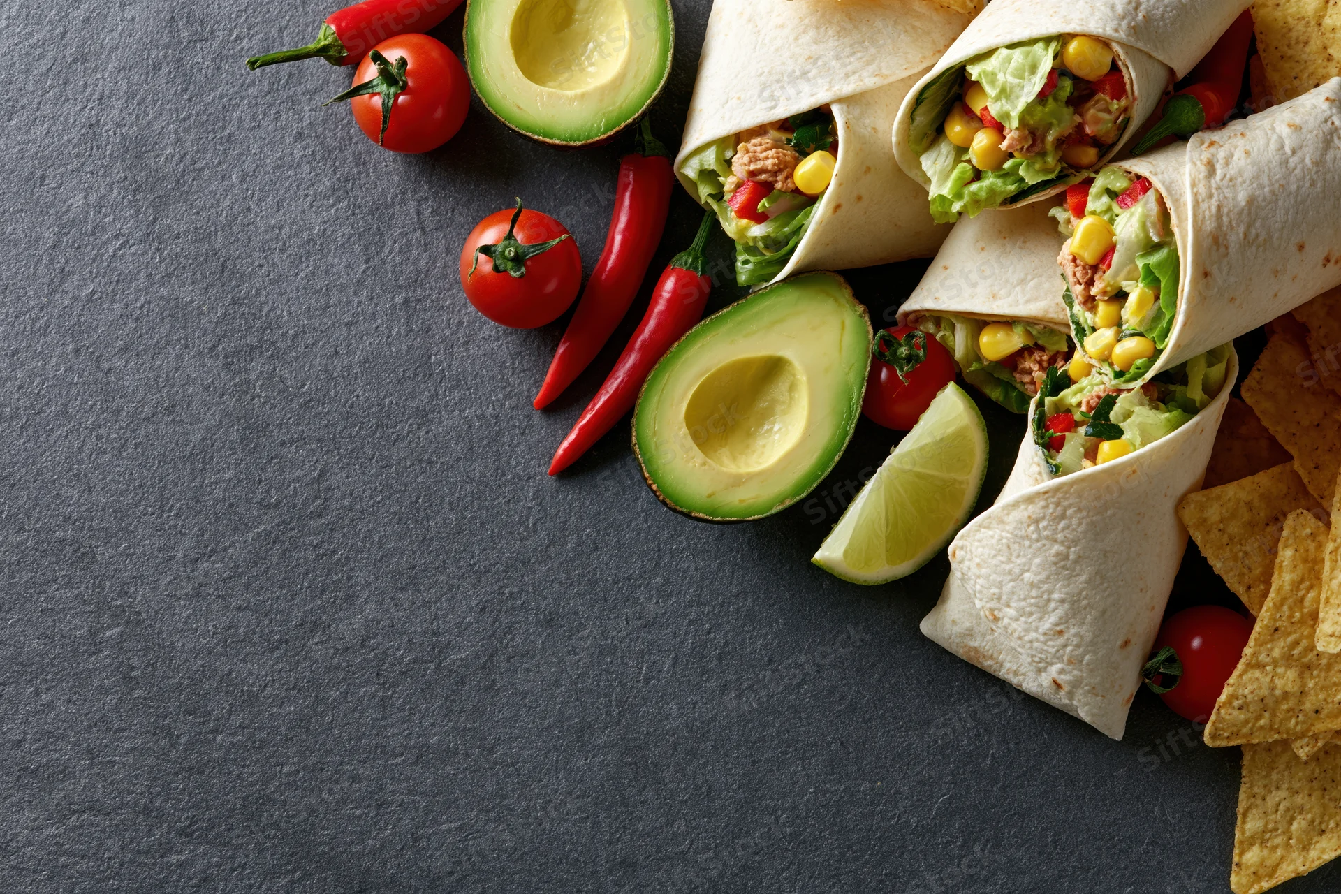 Stuffed tortilla wraps with avocado, tomato, chili, lime, corn, and meat arranged on dark slate surface with copyspace