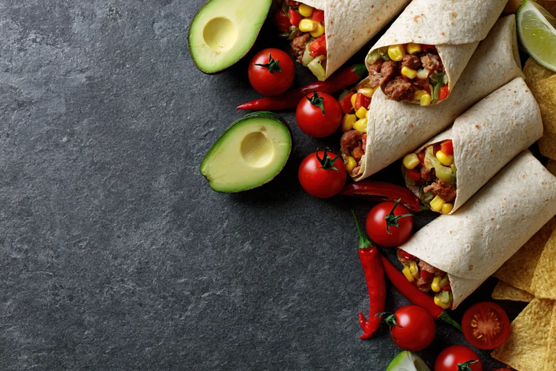 Stuffed tortilla wraps with meat and corn arranged with avocado, tomatoes, chili, lime, and chips on dark stone surface