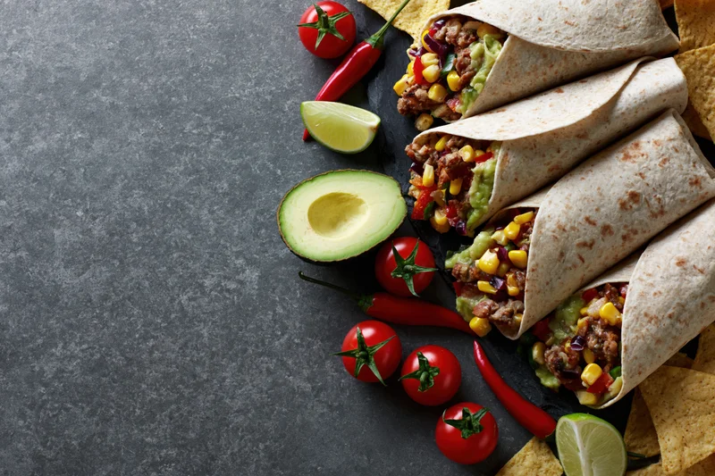 Stuffed tortilla wraps with meat and corn arranged on a dark stone surface with avocado, tomatoes, chili peppers, lime, and tortilla chips