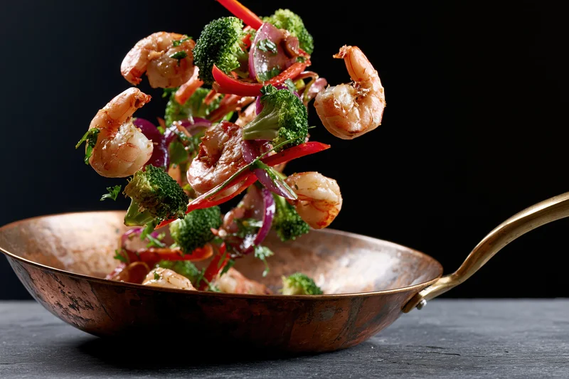 Shrimp and vegetables midair above a copper wok on a dark surface