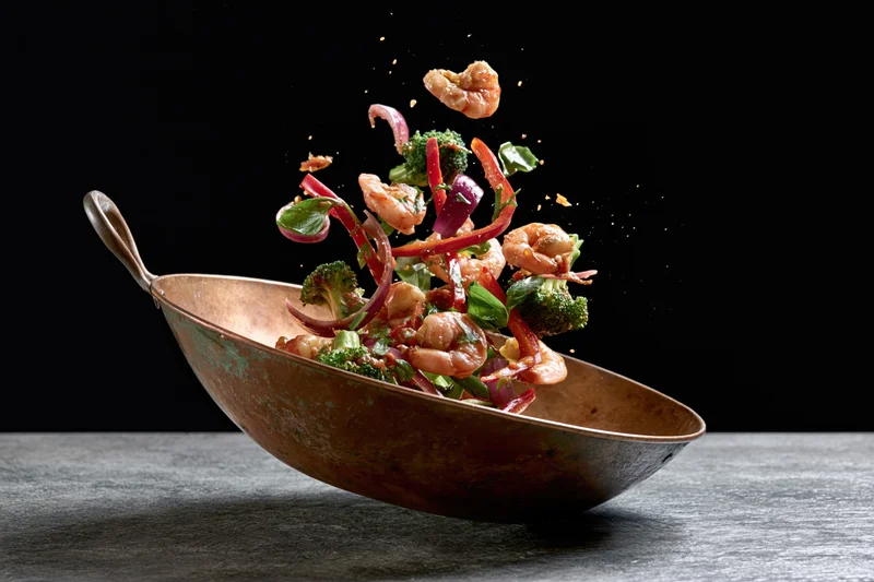 Shrimp and vegetable stir-fry ingredients flying mid-air above a copper wok on a dark surface