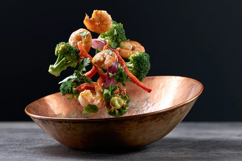 Shrimp and vegetables falling into a copper bowl midair