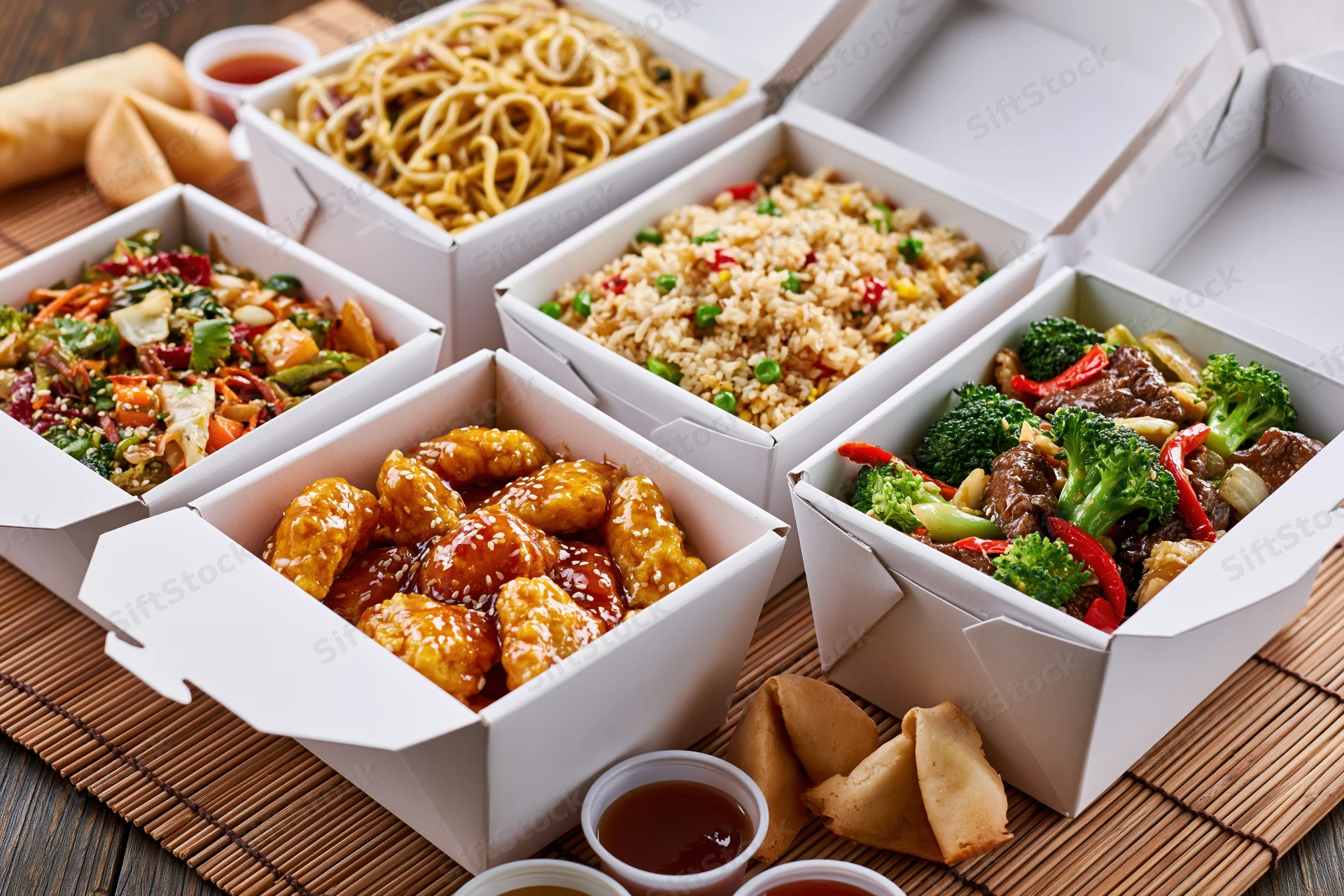 Six open white takeout boxes filled with various chinese dishes arranged on a bamboo mat