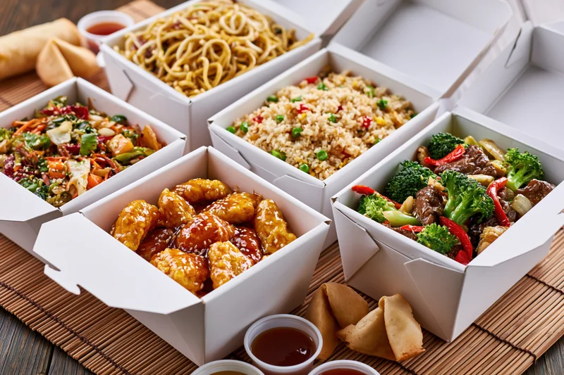 Six open white takeout boxes filled with various chinese dishes arranged on a bamboo mat