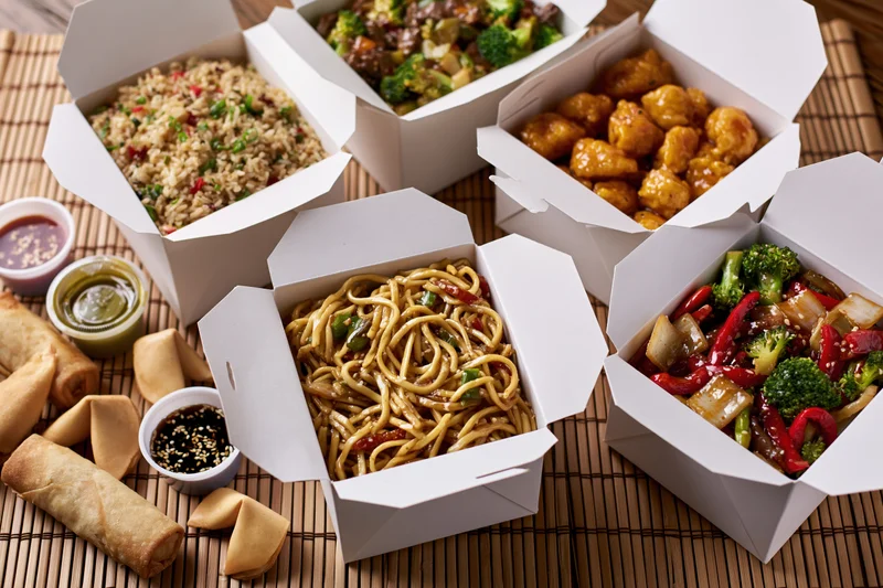 Open white takeout boxes filled with various chinese dishes arranged on a bamboo mat