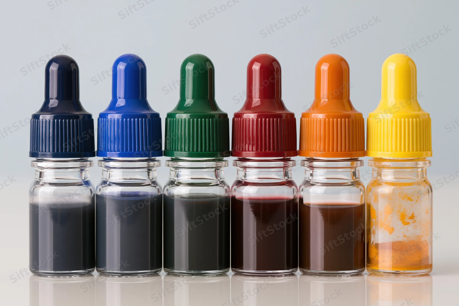 Six glass dropper bottles filled with colored liquid arranged in a row on a reflective white surface