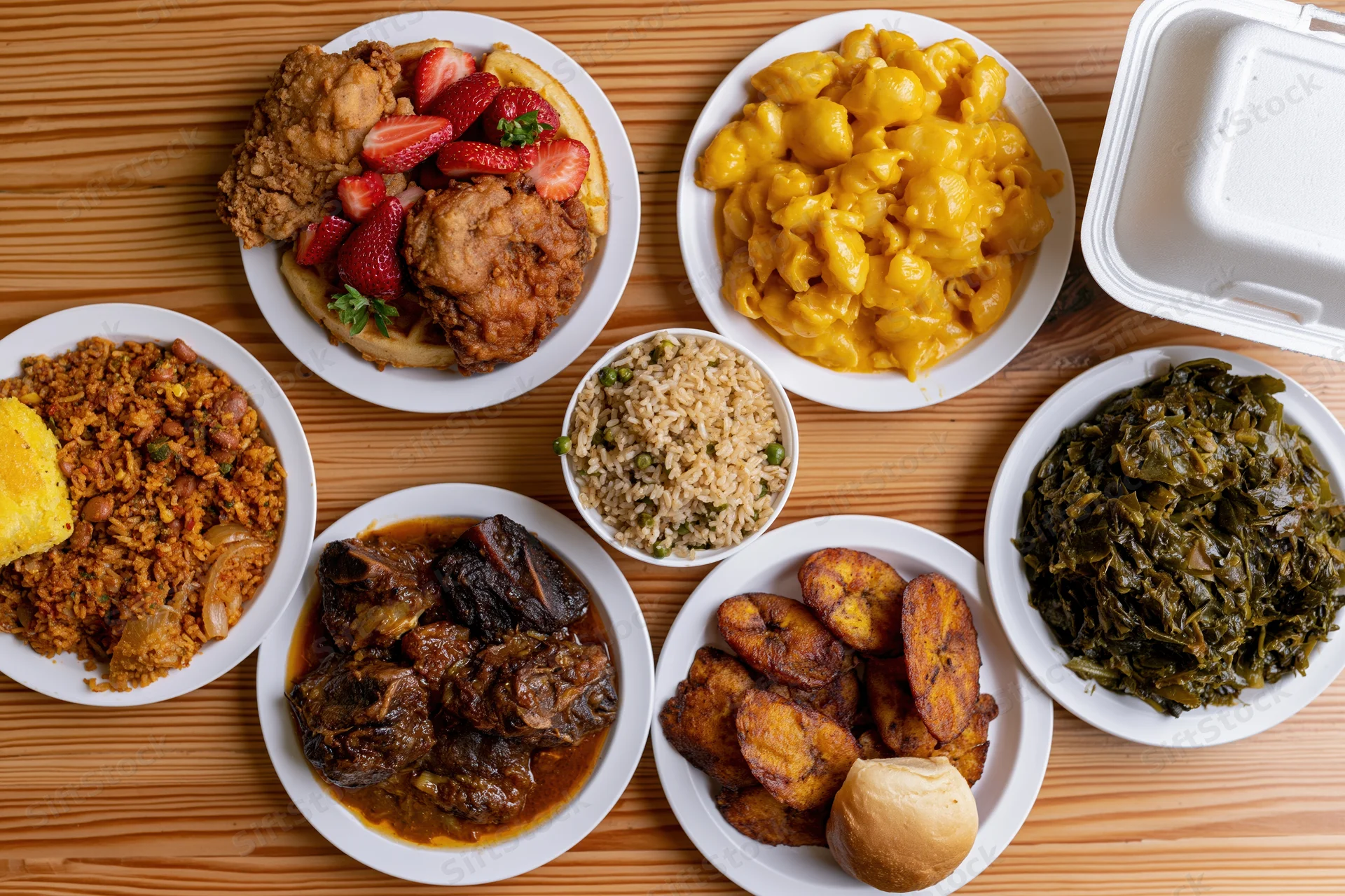 Various dishes including fried chicken, mac and cheese, collard greens, and rice served on white plates and a foam container on a wooden table