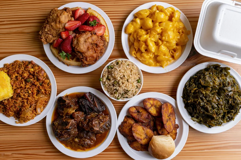 Various dishes including fried chicken, mac and cheese, collard greens, and rice served on white plates and a foam container on a wooden table