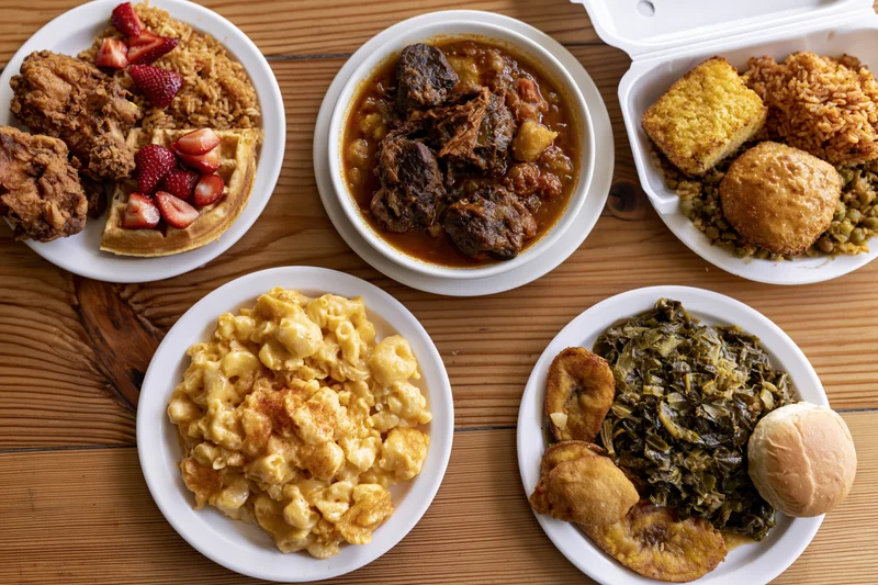 Soul food dishes arranged on wooden table with plates and takeout container