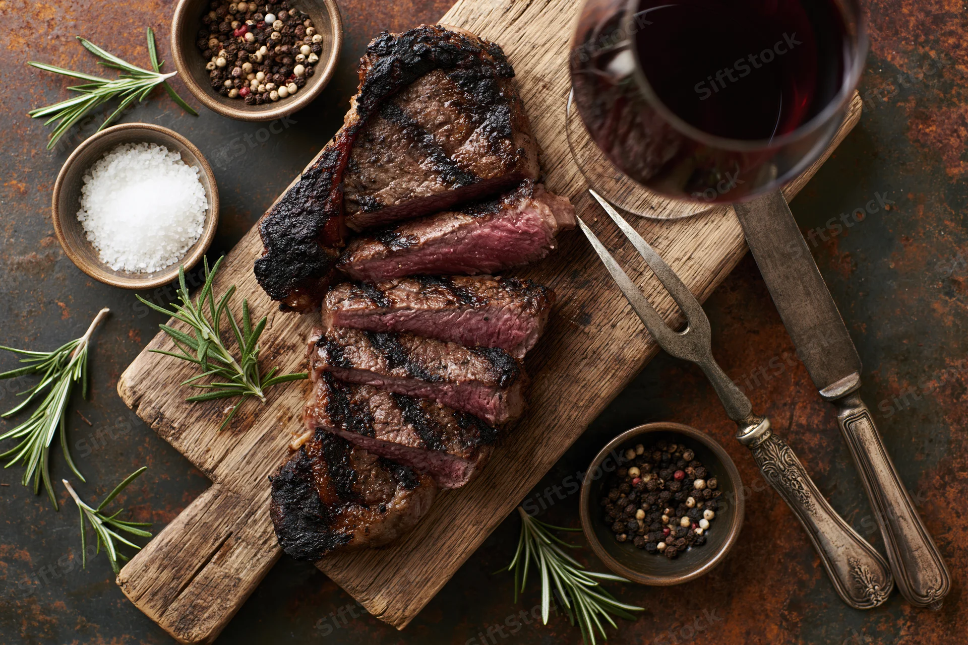 Sliced grilled ribeye steak on a wooden cutting board with herbs, spices, wine and silverware arranged around it for dinner
