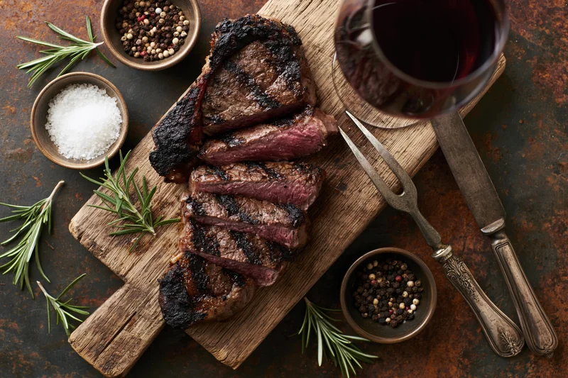 Sliced grilled ribeye steak on a wooden cutting board with herbs, spices, wine and silverware arranged around it for dinner