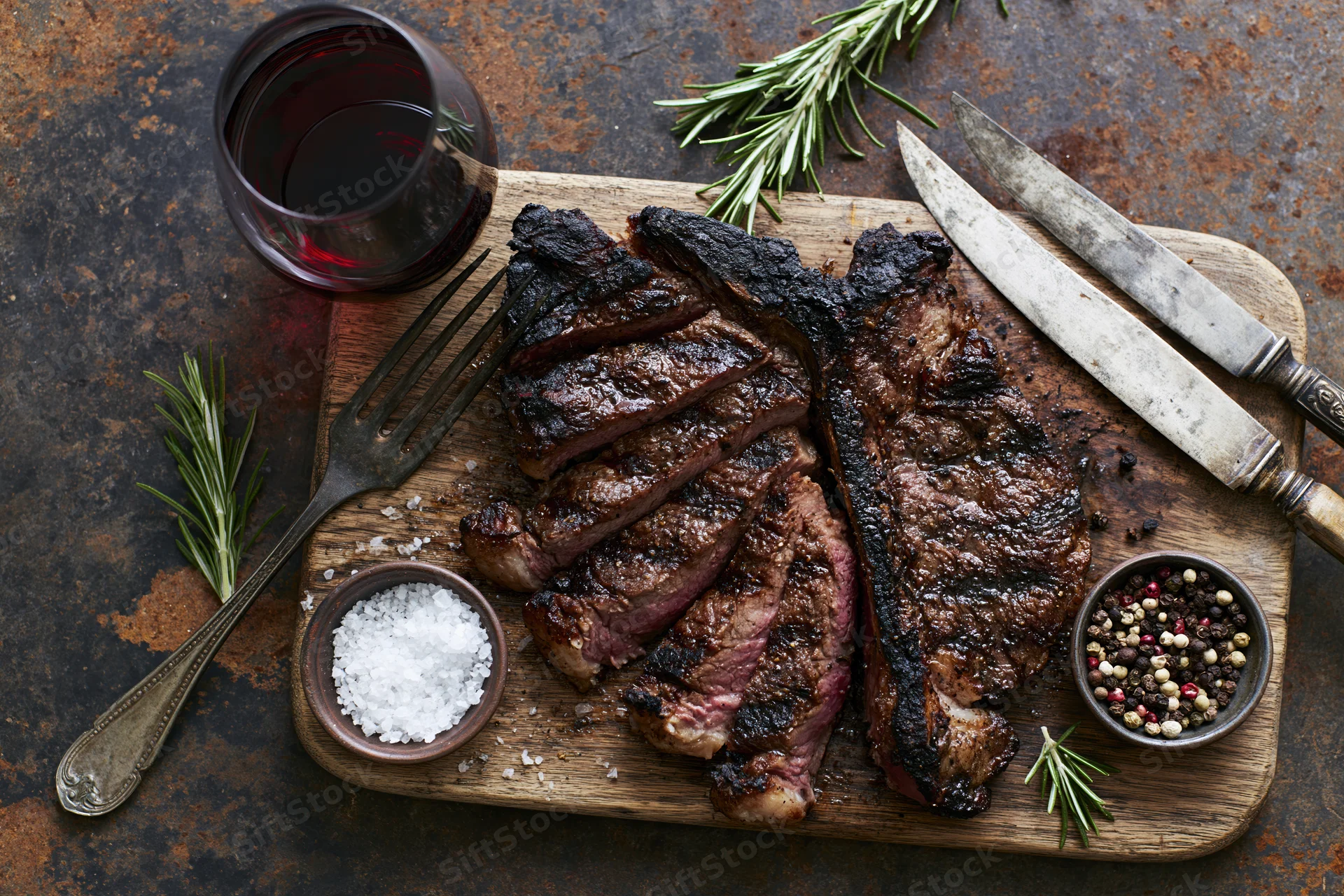 Sliced grilled ribeye steak on a wooden cutting board with herbs, salt, pepper, wine, and utensils