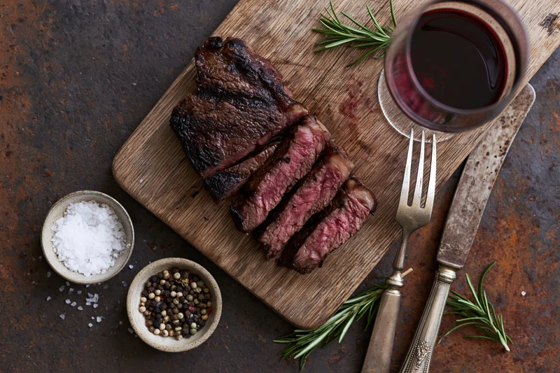 Sliced grilled ribeye steak on a wooden cutting board with wine, herbs, and seasonings on a dark textured surface