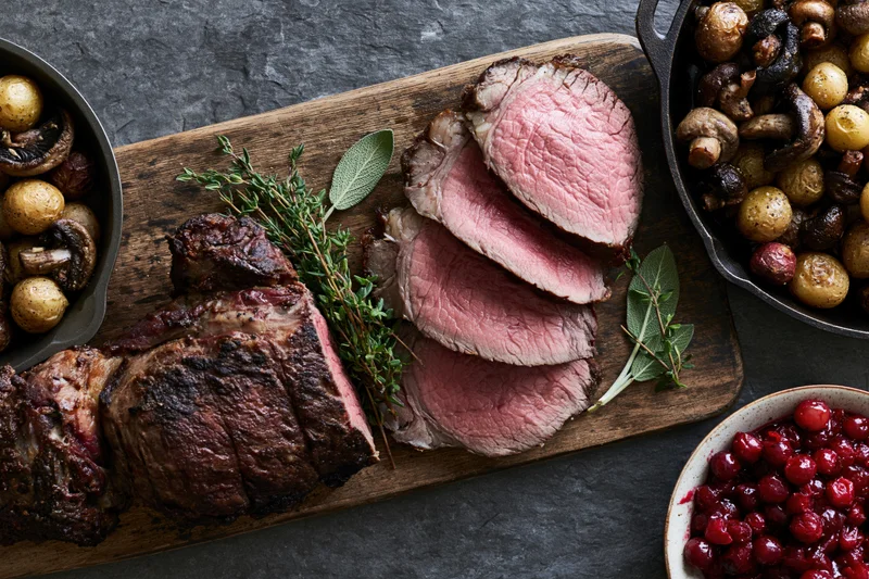 Sliced roast beef on a wooden board with herbs and side dishes arranged for serving