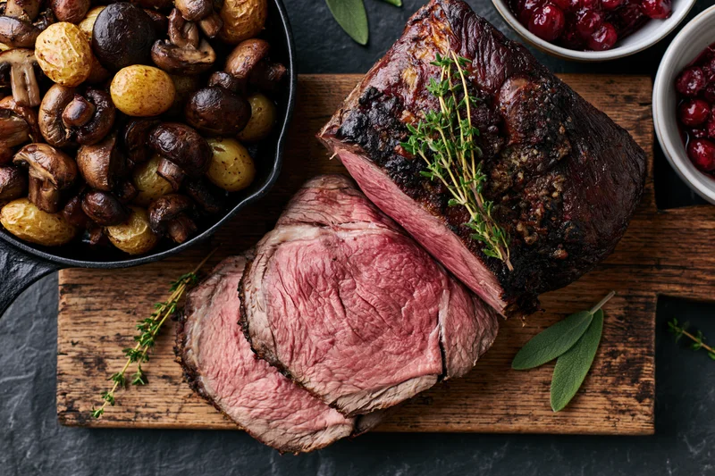 Sliced roast beef on a wooden cutting board with thyme and cranberry sauce beside a skillet of mushrooms and potatoes