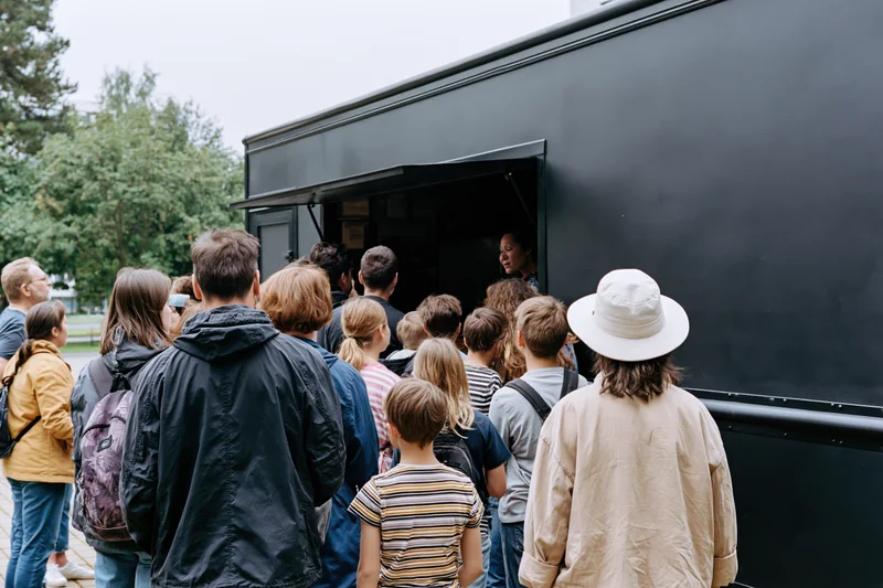 Crowd of adults and children waiting in line at a black food truck's serving window outdoors