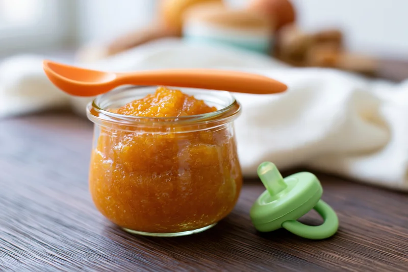Small glass jar filled with orange baby food puree on wooden table with spoon and pacifier nearby