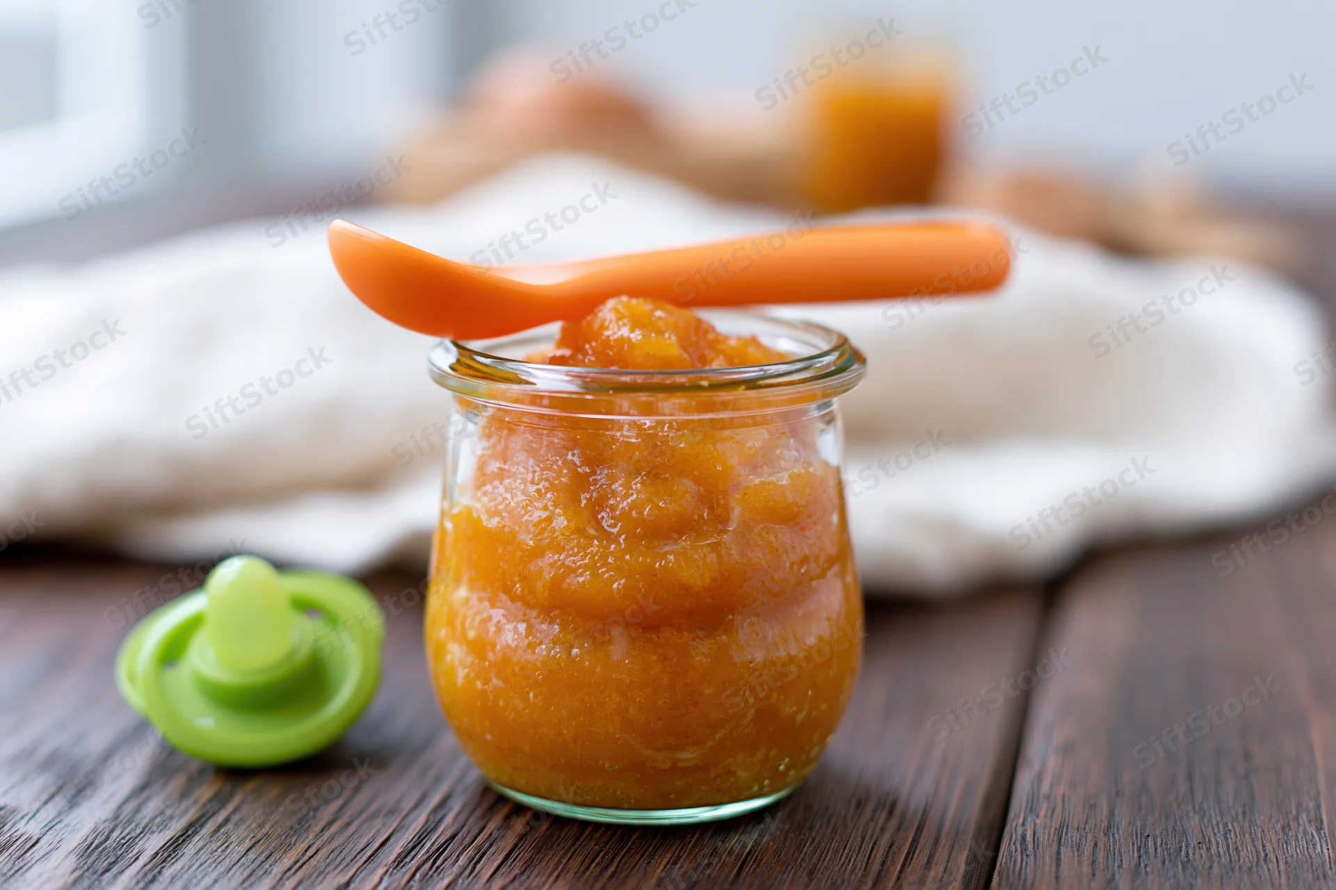 Small glass jar filled with orange baby food puree on wooden table with spoon and pacifier nearby