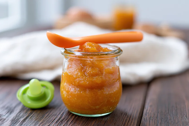 Small glass jar filled with orange baby food puree on wooden table with spoon and pacifier nearby