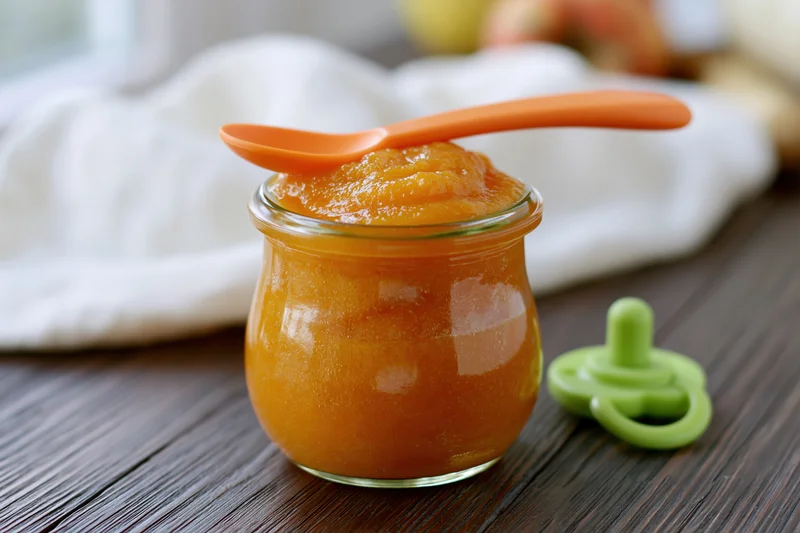 Small glass jar of orange baby food puree with spoon and pacifier on wooden table