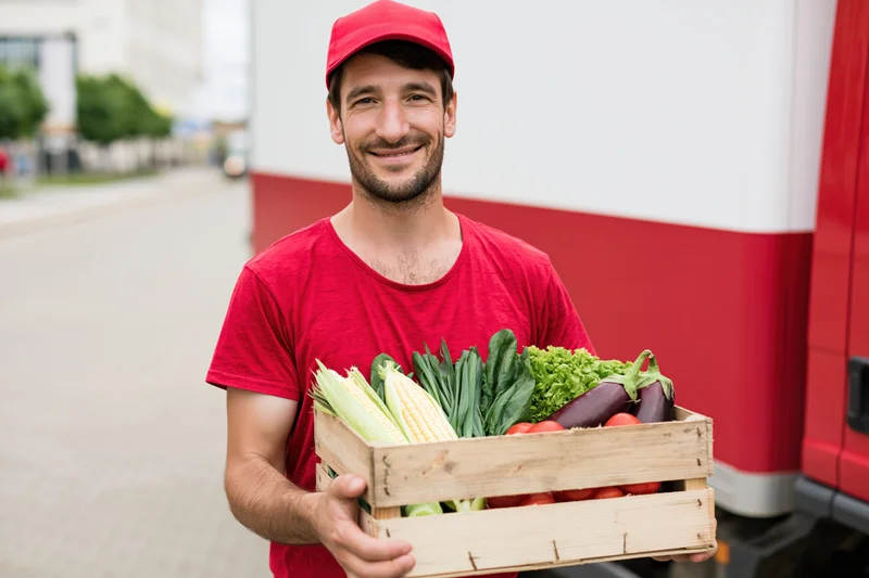 Smiling delivery worker holding wooden crate of fresh vegetables outdoors near delivery truck