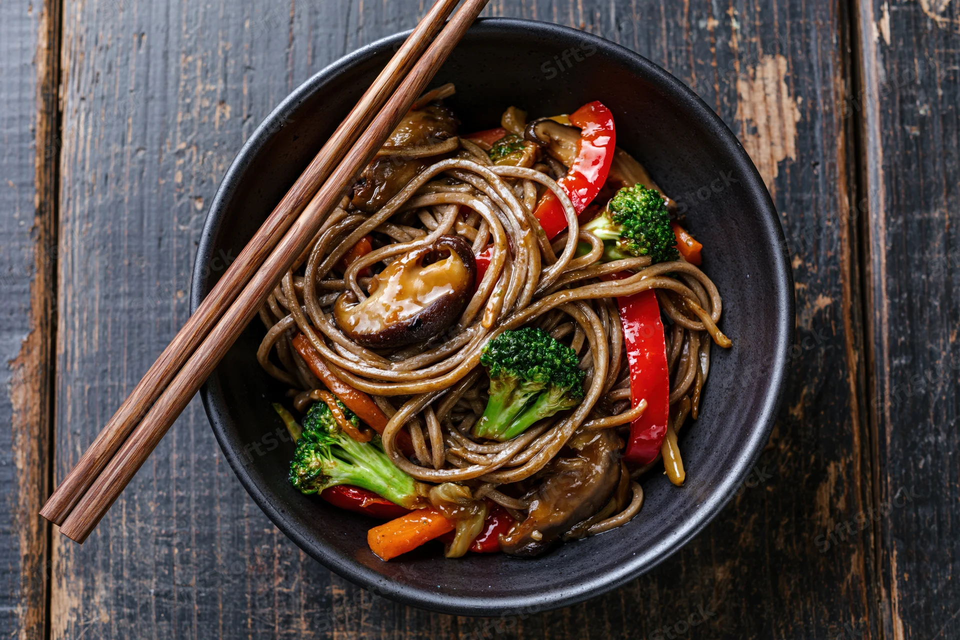 Soba noodle stir-fry with shiitake mushrooms, broccoli, and bell pepper in a black bowl on a dark wooden table, served with chopsticks