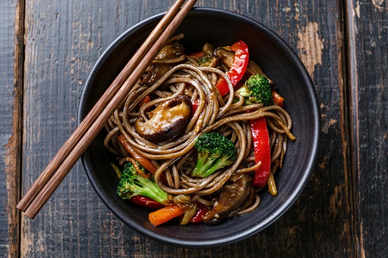 Soba noodle stir-fry with shiitake mushrooms, broccoli, and bell pepper in a black bowl on a dark wooden table, served with chopsticks