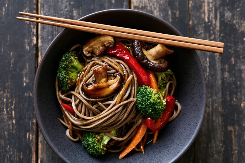 Soba noodle stir fry with shiitake mushrooms broccoli and red pepper in a dark bowl on wooden table with chopsticks resting on top