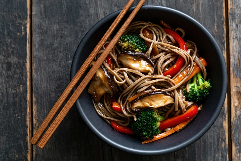 Soba noodle stir fry with shiitake mushrooms broccoli and red pepper in a black bowl with chopsticks on dark wooden table