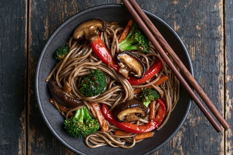 Soba noodles stir-fried with shiitake mushrooms and broccoli in a dark bowl with chopsticks on a wooden table