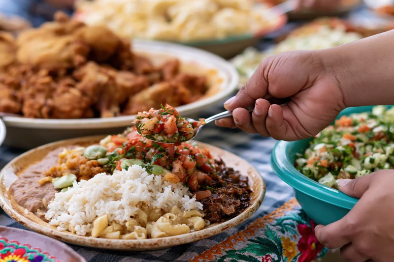 Hand spooning fresh pico de gallo salsa onto a plate with rice, beans, and meat at a festive meal