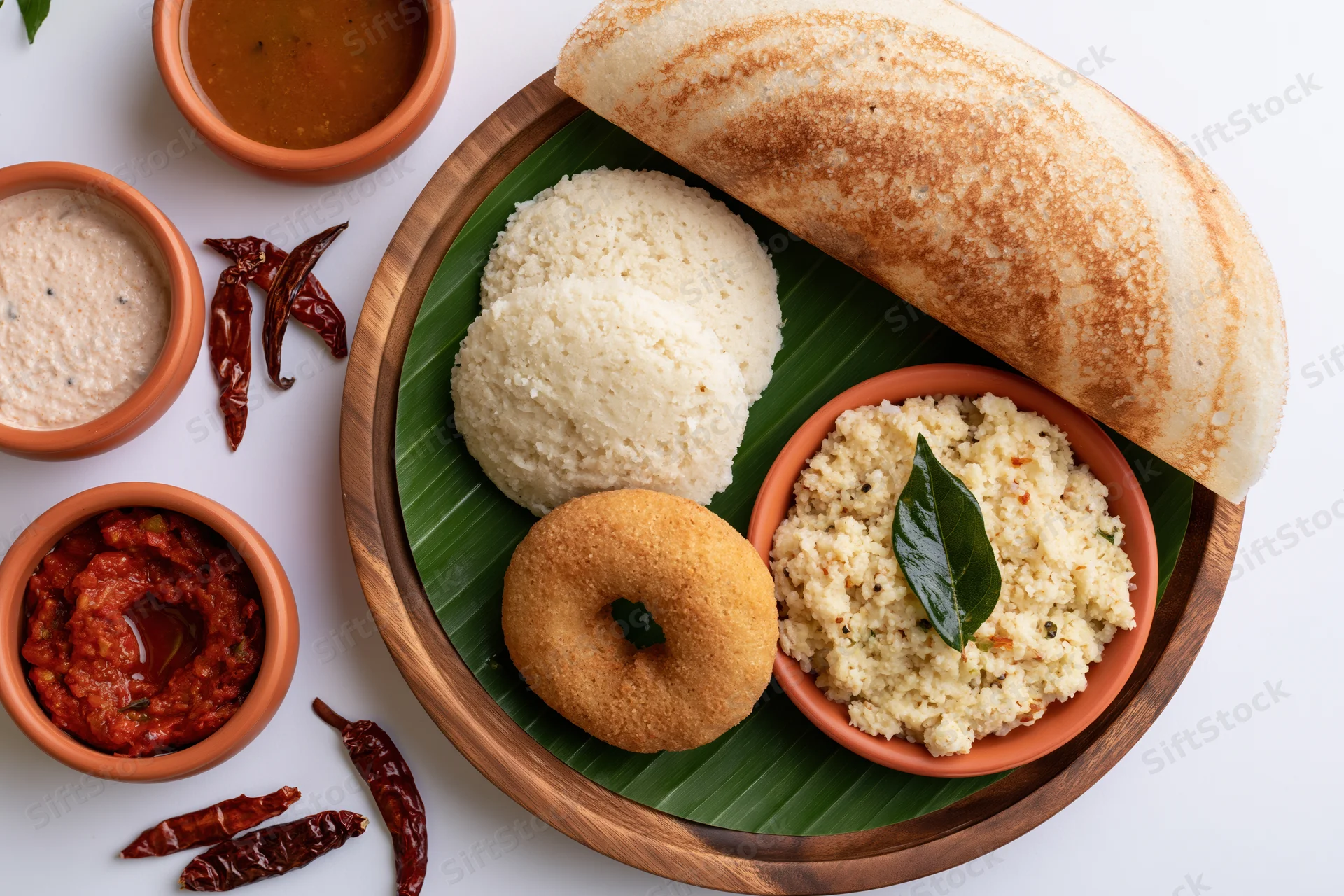 South indian breakfast spread with dosa, idli, vada, sambar and chutney on a wooden tray lined with banana leaf