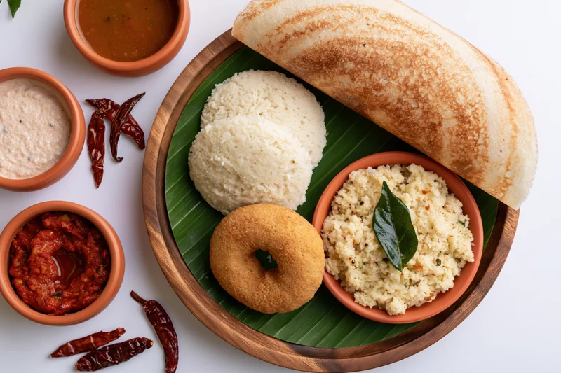 South indian breakfast spread with dosa, idli, vada, sambar and chutney on a wooden tray lined with banana leaf