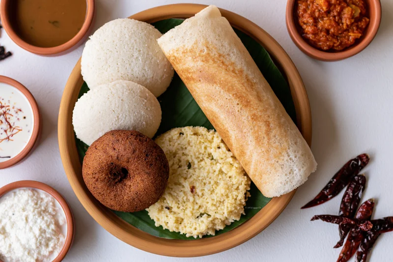 South indian breakfast spread with idli, dosa, vada and upma on a wooden tray with chutney and sambar bowls arranged overhead
