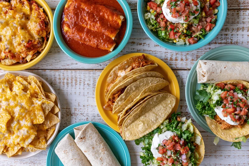 Spread of mexican dishes including tacos, burritos, nachos and enchiladas on a whitewashed wood table