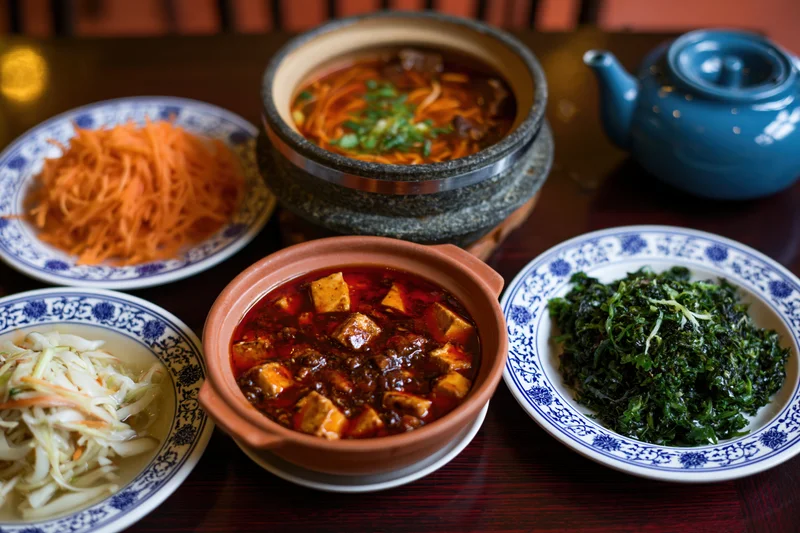 Traditional chinese dishes arranged on dark wooden table including mapo tofu, noodles, vegetables, and tea