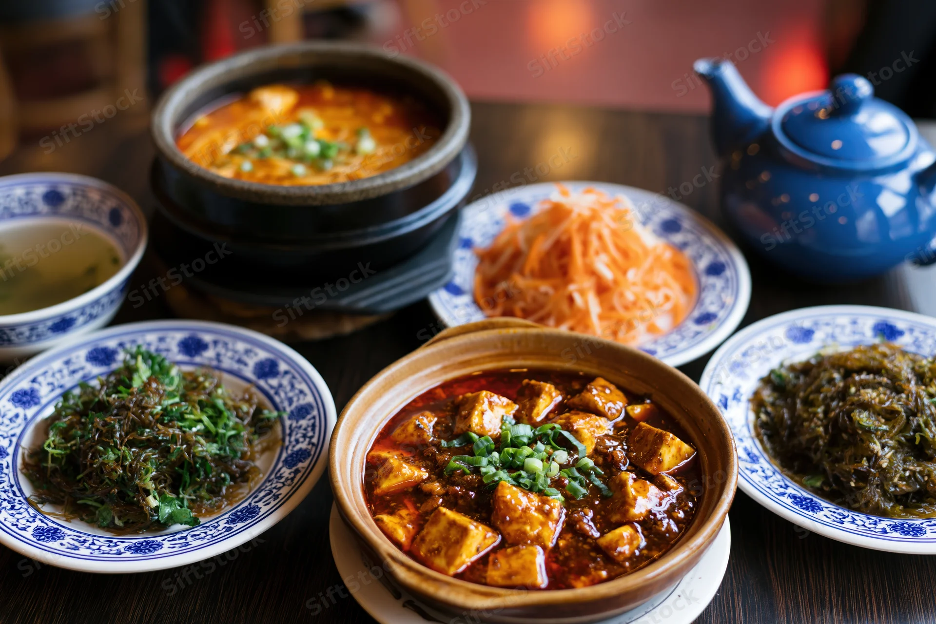 Traditional chinese dishes arranged on dark wooden table including mapo tofu, soup, pickled radish, seaweed salad, and blue porcelain bowls with teapot