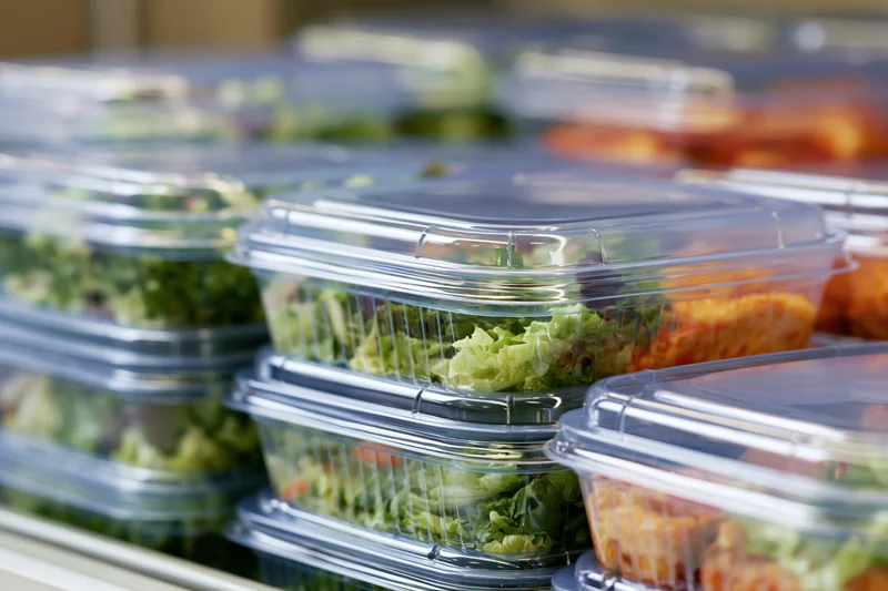Stacked clear plastic clamshell containers filled with salads and meals on a refrigerated shelf