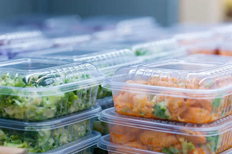Stacked clear plastic clamshell containers filled with salads and chicken meals arranged in rows on a kitchen counter