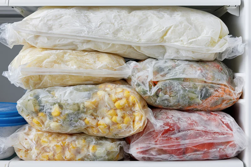 Stacked plastic bags of frozen vegetables inside a white freezer shelf